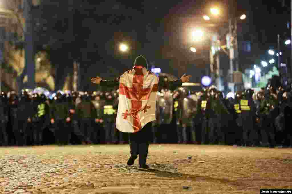 A protester wearing the Georgian flag walks toward a line of riot police outside the Georgian parliament building on March 7. Violent protests broke out after legislators gave preliminary backing to a bill that would require organizations operating in Georgia that receive more than 20 percent of their funding from abroad to register as a &quot;foreign agent.&quot;&nbsp;&nbsp;